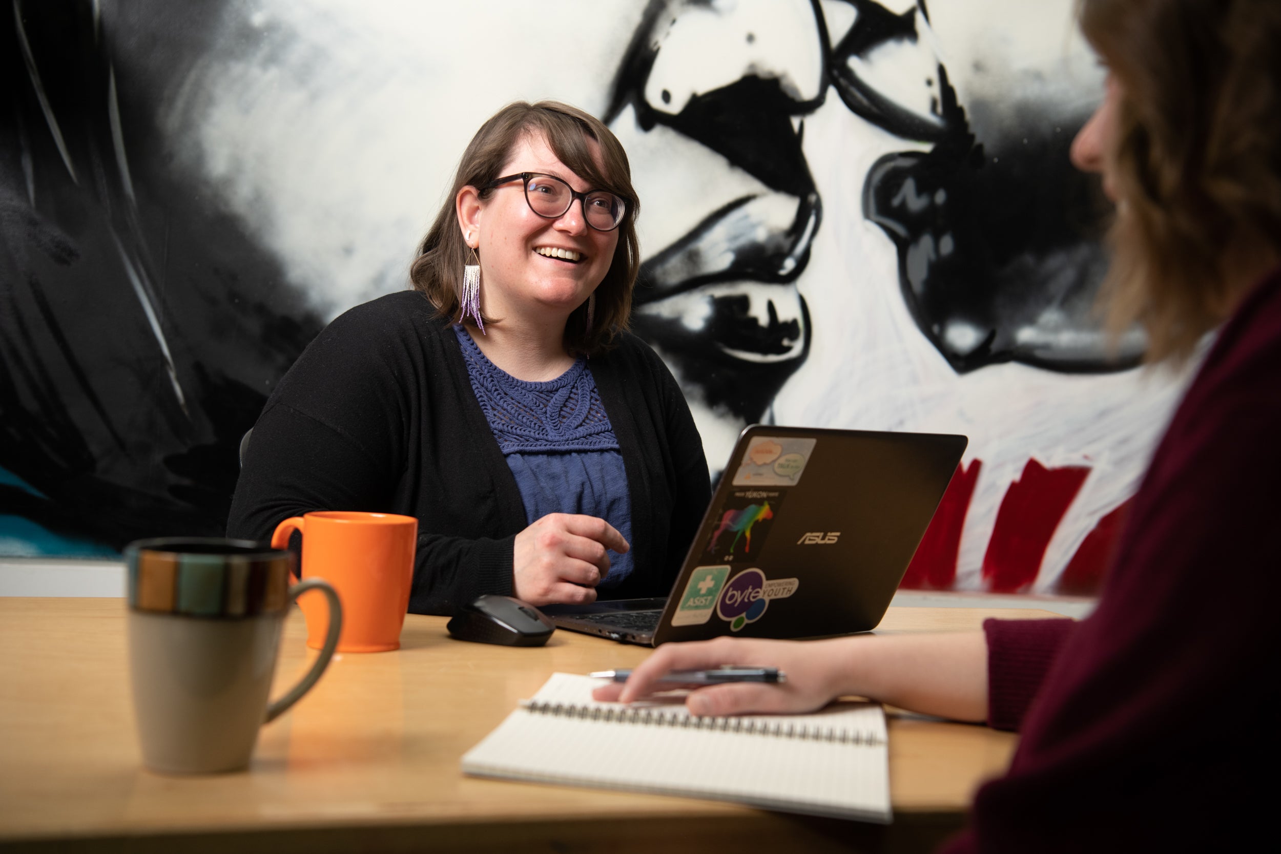Josi Leideritz sitting at a desk in front of her laptop, smiling. In the foreground, the silhouette of a person and their hand is visible holding a pen