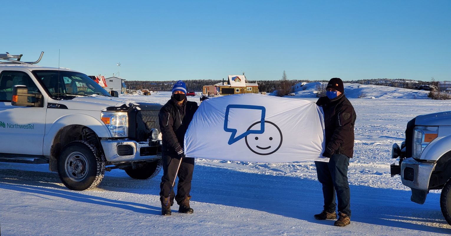 Neil and Tim are standing on Great Slave Lake, NT holding a #BellLetsTalk Day flag to show support for people living with mental health struggles in the north. There is a winter ice road that connects Yellowknife to the small village of Dettah.]