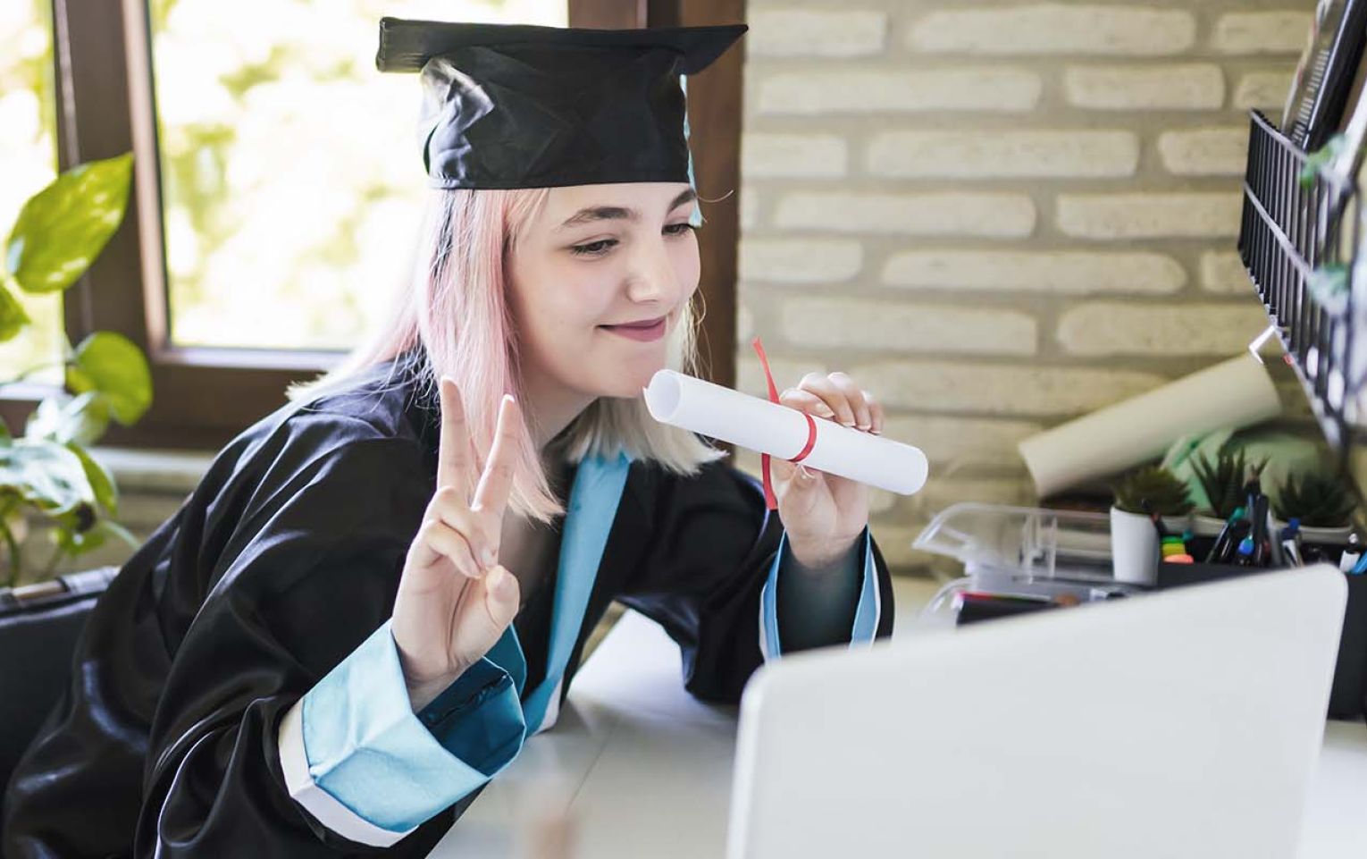 Northern stars shining bright: Congratulations to our 2022 Northern Futures scholarship winners A young lady with light pink hair dressed in a graduation robe, sitting in front of a laptop. She is smiling, holding what appears to be a rolled up diploma. Her other hand is holding up her pointer and middle fingers - commonly referred to as the peace sign. In the background is a window lighting up the room.