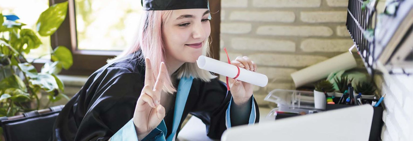 Northern stars shining bright: Congratulations to our 2022 Northern Futures scholarship winners A young lady with light pink hair dressed in a graduation robe, sitting in front of a laptop. She is smiling, holding what appears to be a rolled up diploma. Her other hand is holding up her pointer and middle fingers - commonly referred to as the peace sign. In the background is a window lighting up the room.
