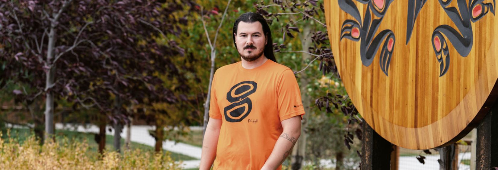 Kailen Gingall wears his Kinghą̈r Orange Shirt Kailen Gingall stands in front of a largen wooden crest at the Kwanlin Dun Cultural Centre. He is wearing the orange shirt he designed.