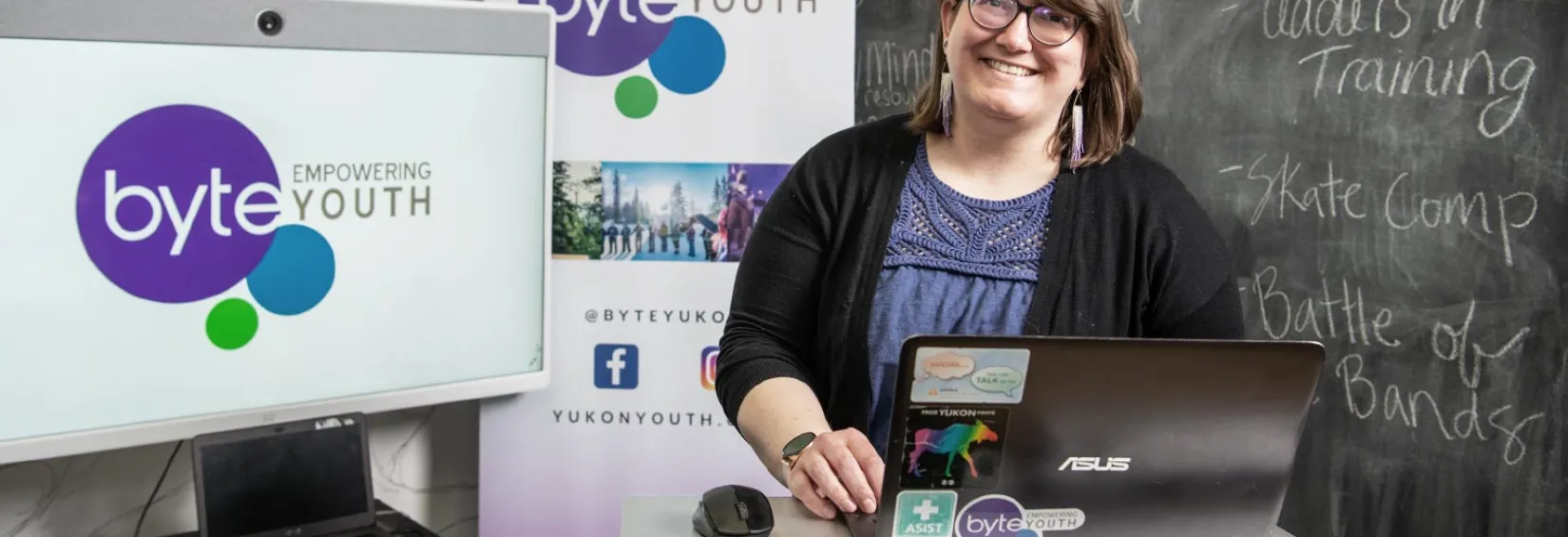Josi Leideritz, BYTE's executive director Josi Leideritz, BYTE's executive director is at a standing desk with a laptop. Behind are a chalkboard with various session titles: Leaders In Training, Skate Camp, and Battle of the bands. On the other side of her is a screen and a pop up banner, both showing the byte logo.