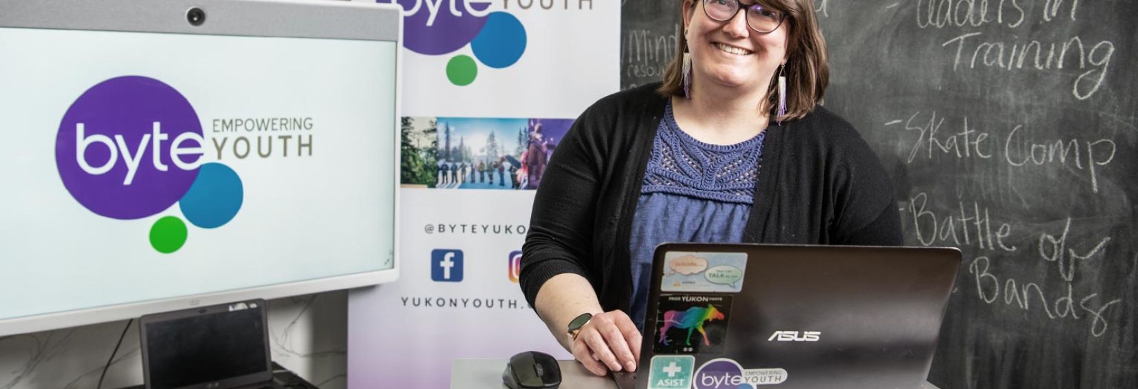 Josi Leideritz, BYTE's executive director is at a standing desk with a laptop. Behind are a chalkboard with various session titles: Leaders In Training, Skate Camp, and Battle of the bands. On the other side of her is a screen and a pop up banner, both showing the byte logo. 