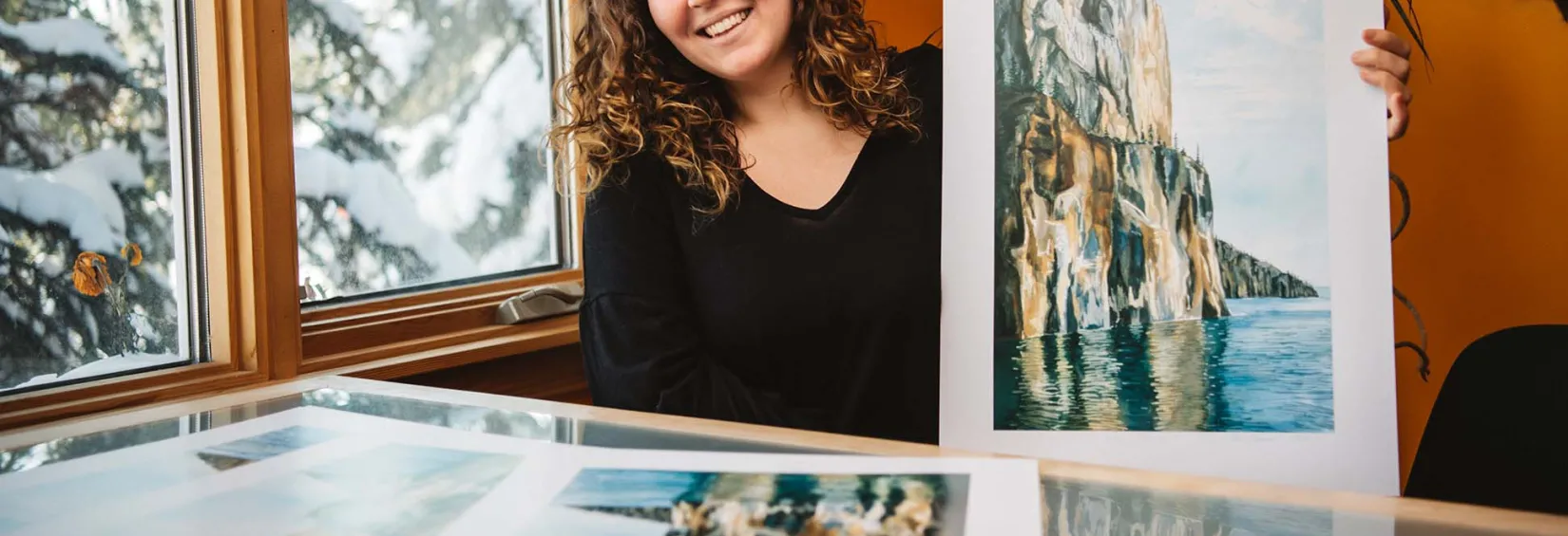 Northwest Territories directory art winner showcases the great cliffs of Etthen Island Maggie Davies, Northwest Territories Directory Art Contest winner, sits at a desk and smiles while holding a print of her art. She is next to a window and snowy trees are visible outside. Inside, the room has a nice warm hue.