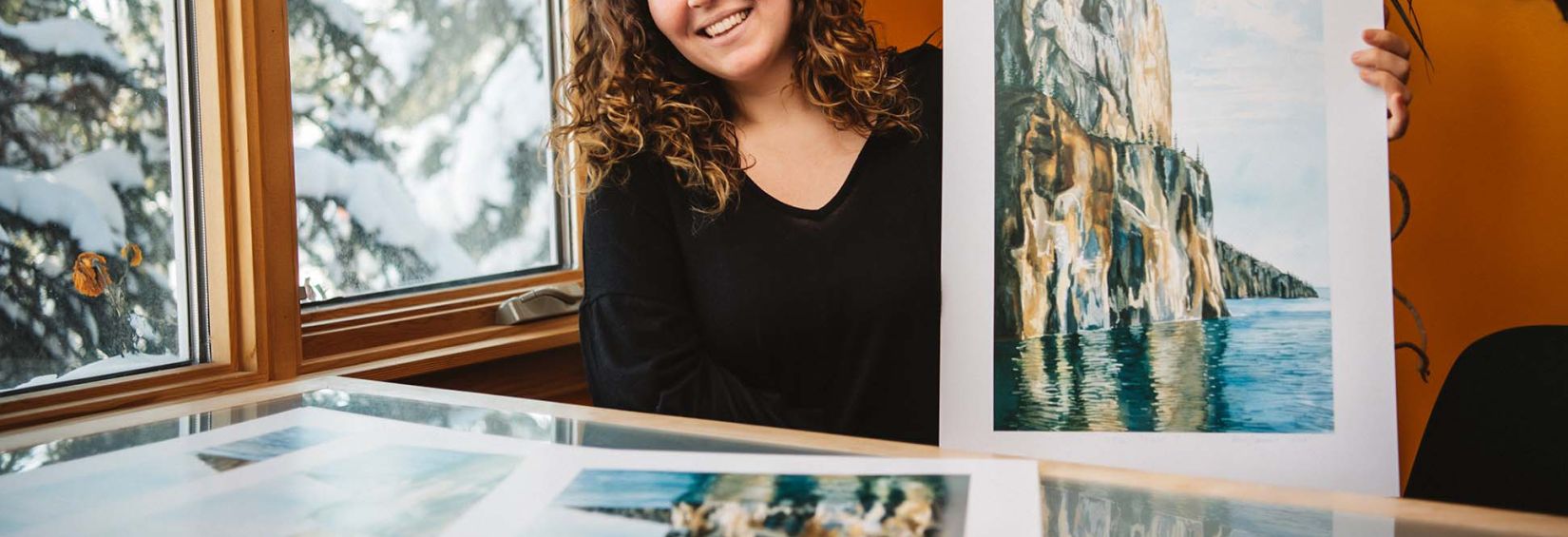 Northwest Territories directory art winner showcases the great cliffs of Etthen Island Maggie Davies, Northwest Territories Directory Art Contest winner, sits at a desk and smiles while holding a print of her art. She is next to a window and snowy trees are visible outside. Inside, the room has a nice warm hue.