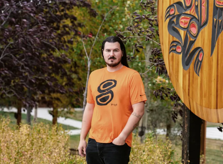 Kailen Gingall wears his Kinghą̈r Orange Shirt Kailen Gingall stands in front of a largen wooden crest at the Kwanlin Dun Cultural Centre. He is wearing the orange shirt he designed.